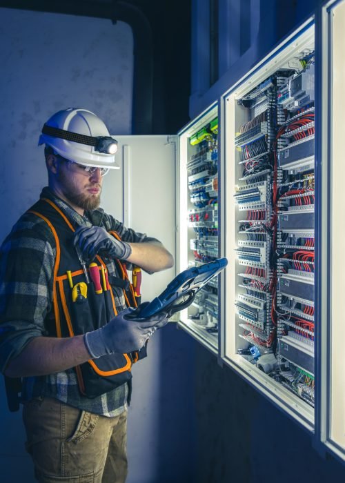 Electrician focused on work in switchboard with fuses, using tablet. Young adult electrical engineer in special clothes with flashlight on helmet in dark room with emergency lights in background.